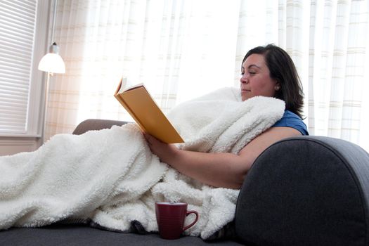 A single woman reads a book, happy to be at home alone with a blanket and warm drink 