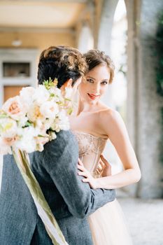 Groom hugs bride with a bouquet of flowers on an old terrace with arches entwined with green ivy. Lake Como. Close up. High quality photo