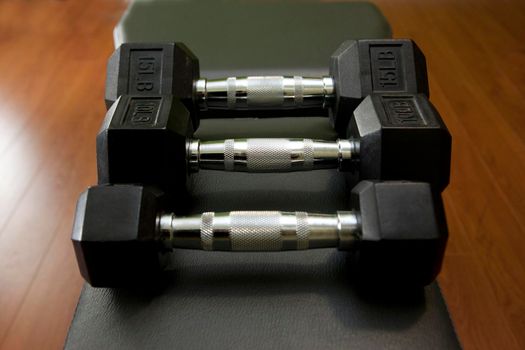 Three small weight dumbells on a weight bench with a dumbell rack in background 
