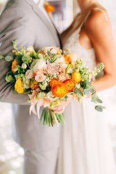 The bride and groom stand hugging and hold a wedding bouquet with orange buttercups, roses and eucalyptus branches close-up . High quality photo