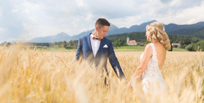 Groom and bride holding hands in wheat field somewhere in Slovenian countryside. Caucasian happy romantic young couple celebrating their marriage.