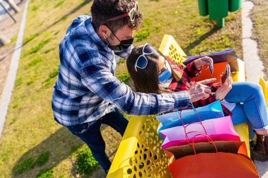 Young heterosexual couple of shopaholic people sitting on a city park bench among colored shopping bags using smartphone and credit card to pay online purchases with new internet mobile connection