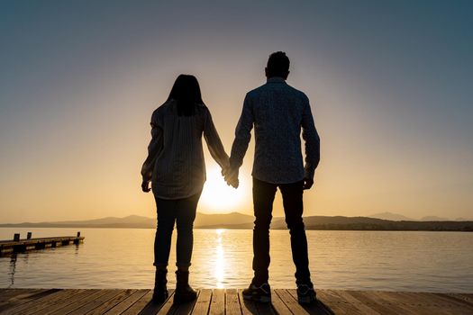 Suggestive silhouette back shot of young couple in love holding hands standing on a pier watching the sun setting or rising in horizon on flat sea ocean water with light reflections. Positive emotion