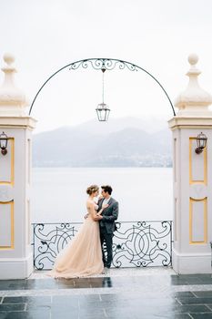 Newlyweds stand in an embrace under an ancient arch leaning on a forged fence against the backdrop of Lake Como. High quality photo