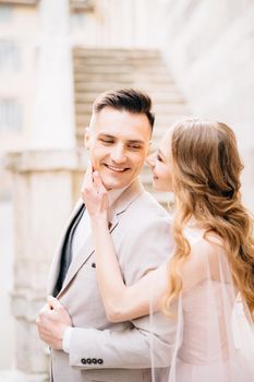 Bride holds her palm on the chin of smiling groom against the background of the steps of an ancient building in Bergamo. High quality photo