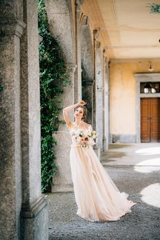 Bride in a beautiful dress with a bouquet of pink flowers stands in the vaulted hall with her hand on her forehead. Lake Como, Italy. High quality photo