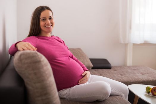 Beautiful pregnant woman enjoys resting on sofa at her home.