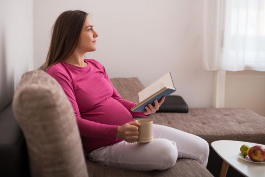 Beautiful pregnant woman enjoys reading book and drinking tea at her home.