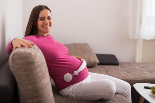 Happy pregnant woman enjoys playing music to her baby while resting at her home.