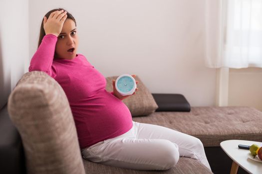 Pregnant woman in panic holding clock while sitting on sofa at her home.