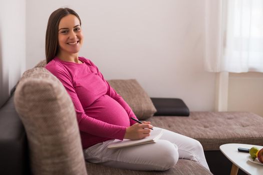 Happy pregnant woman writing to do list in notebook while spending time at her home.