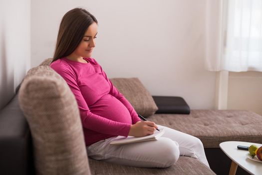 Happy pregnant woman writing to do list in notebook while spending time at her home.