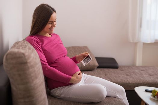 Pregnant woman sitting on the sofa and holding ultrasound picture.