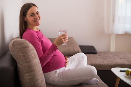 Beautiful pregnant woman enjoys drinking water while  resting on sofa at her home.