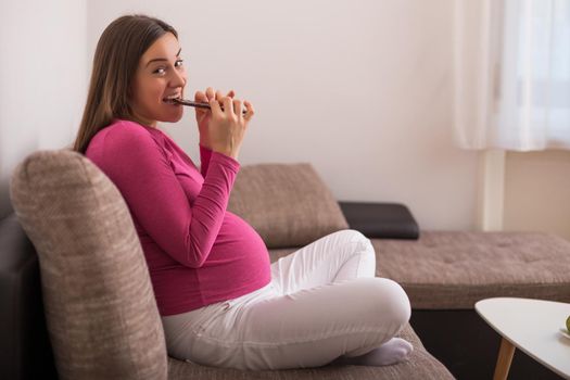 Pregnant woman eating chocolate while sitting on sofa in her home.