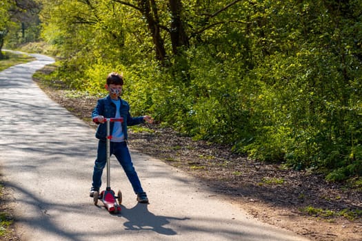 Full-length shot of a boy wearing jeans, a denim jacket and a face mask playing with a scooter in a park on a sunny day