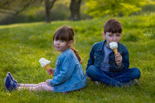 Portrait of a cute boy and a girl wearing blue jackets sitting on a lawn on a sunny day eating ice cream