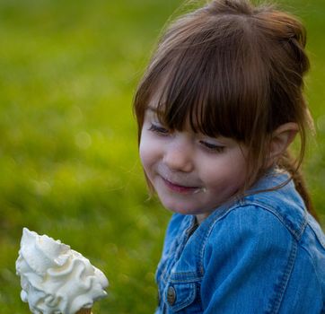 Close-up of a cute, brown-haired, blue-eyed girl, wearing a blue denim jacket eating an ice cream on the grass on a sunny day