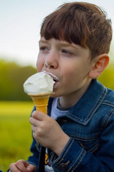 Close-up of a cute, redhead, blue-eyed boy, wearing a blue denim jacket eating an ice cream on the grass on a sunny day