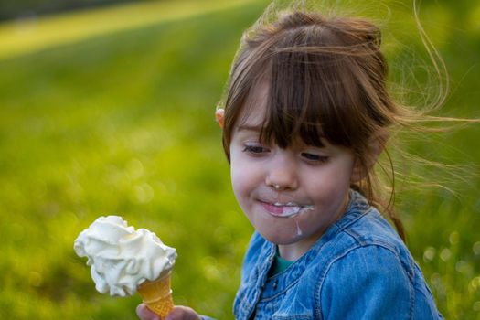 Close-up of a cute, brown-haired, blue-eyed girl, wearing a blue denim jacket eating an ice cream on the grass on a sunny day