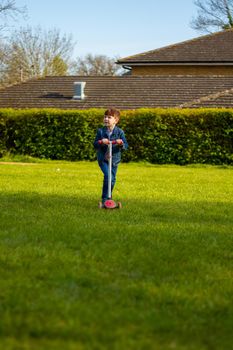 Wide-shot of a cute, redhead boy wearing jeans and a blue denim jacket playing with a scooter in a lawn on a sunny day