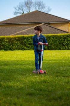 Wide-shot of a cute, redhead boy wearing jeans and a blue denim jacket playing with a scooter in a lawn on a sunny day