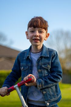 Medium-shot of a cute, redhead boy wearing jeans and a blue denim jacket playing with a scooter in a lawn on a sunny day
