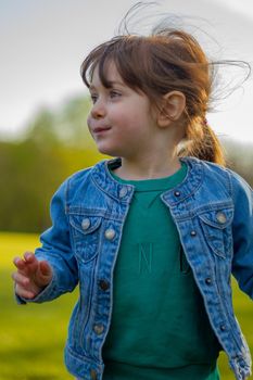 Portrait of a cute, brown-haired, blue-eyed baby girl wearing a blue jacket and green jumper running in a park on a sunny day