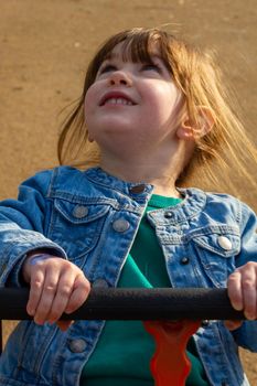 A cute, brown-haired, blue-eyed girl wearing a blue jacket and green jumper sitting in a swing on a sunny spring day