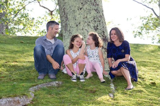 young family sits by the base of a tree outside, laughing and giggling together