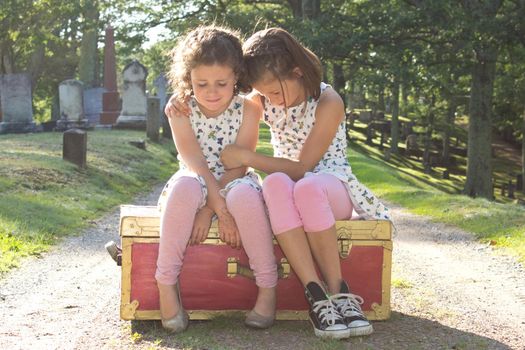 two little girls sit on a suitcase in a cemetery, crying with arms around each other