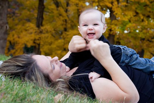 Happy mother playing with her baby girl on some grass with autumn trees in the background.
