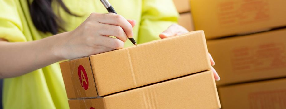 Closeup hand of young asian woman writing address on parcel box at home for delivery order to customer, shipping and logistic, merchant online and seller, business owner or SME, online shopping.