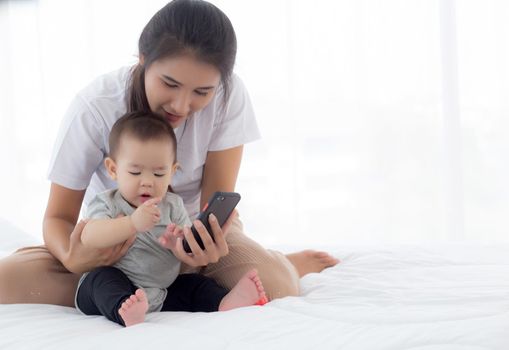 Young asian mother and little baby girl or newborn selfie with smart phone on bed in bedroom, happiness mom and daughter using phone video call at home, two people, family and communication concept.