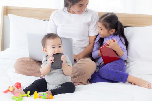 Young asian mother working at home online to internet with laptop computer and daughter looking tablet on bed at bedroom, business woman is freelance and children learning, new normal, family concept.