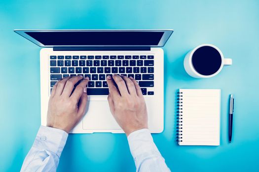 Flat lay, hand of businessman working on laptop computer with cup of coffee on desk in office, workplace and notebook and book on blue background, workspace and copy space, top view, business concept.