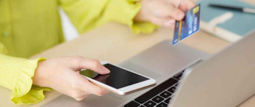 Closeup young asian business woman using smart phone and holding credit card while online shopping and payment with laptop computer on desk at home, female holding debit card, communication concept.