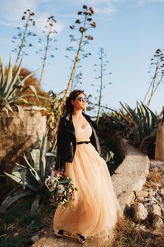 Cute bride in a pastel dress, a leather jacket and sunglasses stands on a path against a background of agaves. High quality photo
