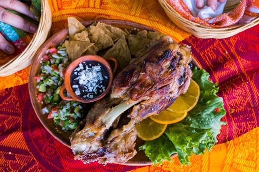Top view of delicious roasted pork with lemon slices, lettuce, tortilla chips, and refried beans on colorful tablecloth. Tasty meat and accompaniments above orange and red table. Mexican style meal