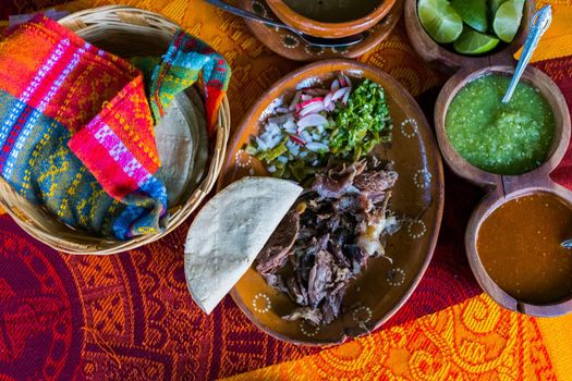 Top view of chopped lamb meat, hot sauces, lime slices, broth, and tortillas on colorful tablecloth. Tasty meal and condiments in clay plates and baskets above red and orange table. Mexican cuisine