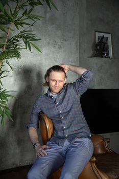 The handsome stylish young man sits on an expensive leather armchair relaxed, hand straightens long curly hair, he is dressed in a blue shirt and trousers, he is looking at the camera. High quality photo