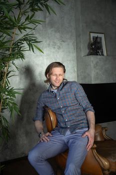 The handsome stylish young man sits on an expensive leather armchair relaxed, long curly hair, he is dressed in a blue shirt and trousers, he is looking at the camera. High quality photo