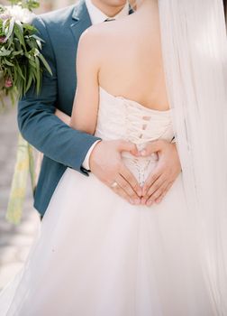 Groom holds his hands in the shape of a heart on the back of the bride in a white dress, hugging her. High quality photo
