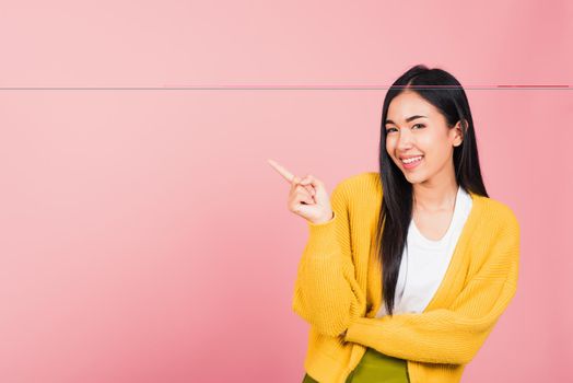 Asian happy portrait beautiful cute young woman standing pointing finger side away presenting product looking to camera, studio shot isolated on pink background with copy space