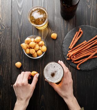 Top view of woman hands holding glass of beer and cheese snack, black wooden background. Flat lay