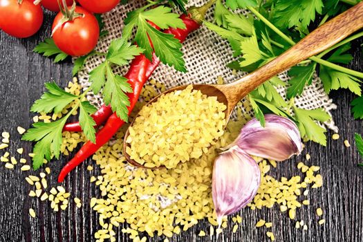 Bulgur groats - steamed wheat grains - in a spoon on sacking, tomatoes, hot peppers, garlic and parsley on a wooden board background from above