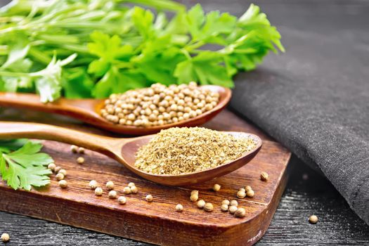 Coriander seeds and ground in two spoons, green fresh cilantro and a napkin on black wooden board background