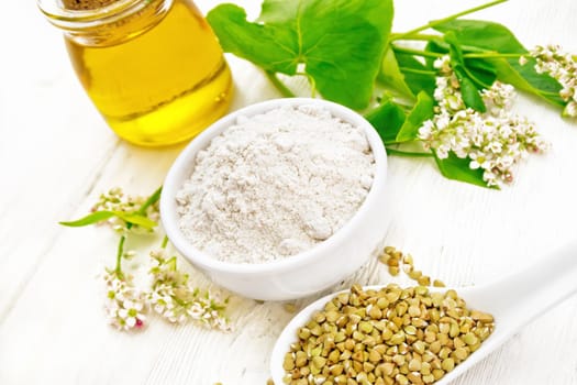 Buckwheat flour from green cereals in a bowl, groats in a spoon and on a table, oil in a glass jar, fresh flowers and buckwheat leaves on a white wooden board background