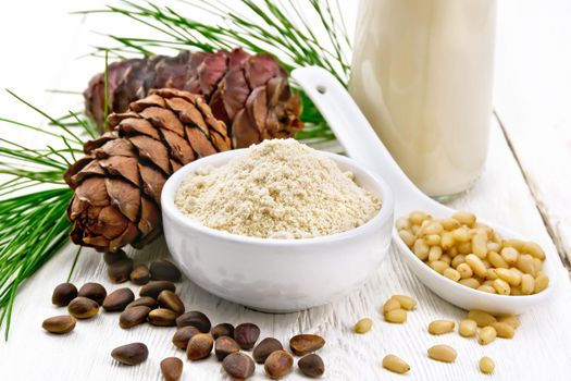 Cedar flour in a bowl, nuts and cones, a spoon with peeled nuts, green pine branch and cedar milk in a bottle on wooden board background