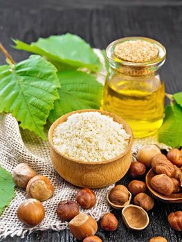 Hazelnut flour in a bowl, nuts in a spoon, oil in glass jar and filbert branch with green leaves on burlap on dark wooden board background